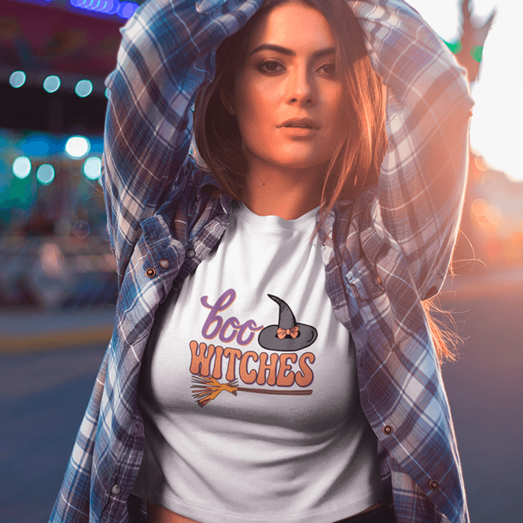 Woman wearing a 'boo witches' t-shirt with a plaid shirt over it, standing in front of a blurred amusement park background.