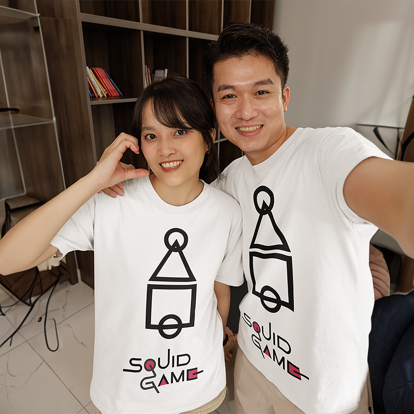 A happy man and woman taking a selfie while wearing matching white 'Squid Game' T-shirts with black geometric symbols. Perfect for couples or themed apparel. Made by Your iDeas Shop