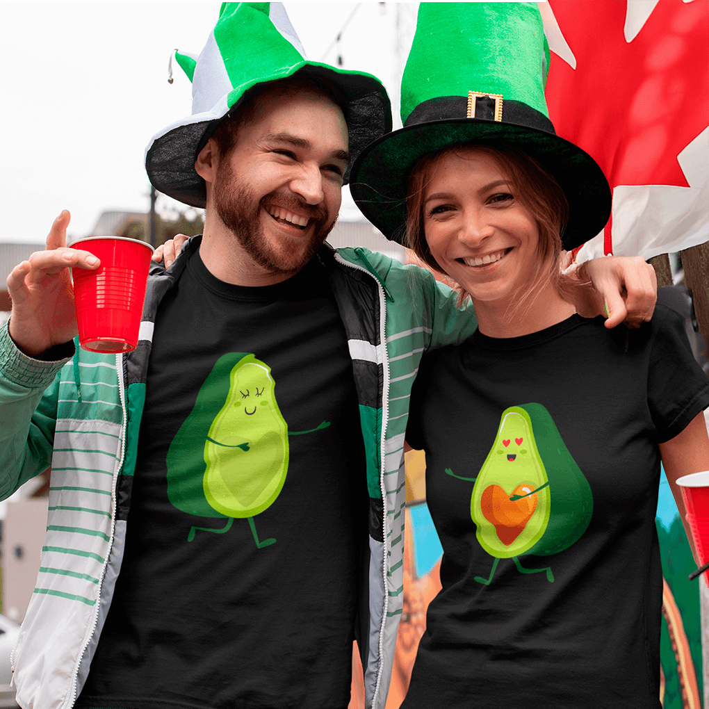 Two people wearing black t-shirts with avocado designs, smiling and posing in front of a colorful flag.