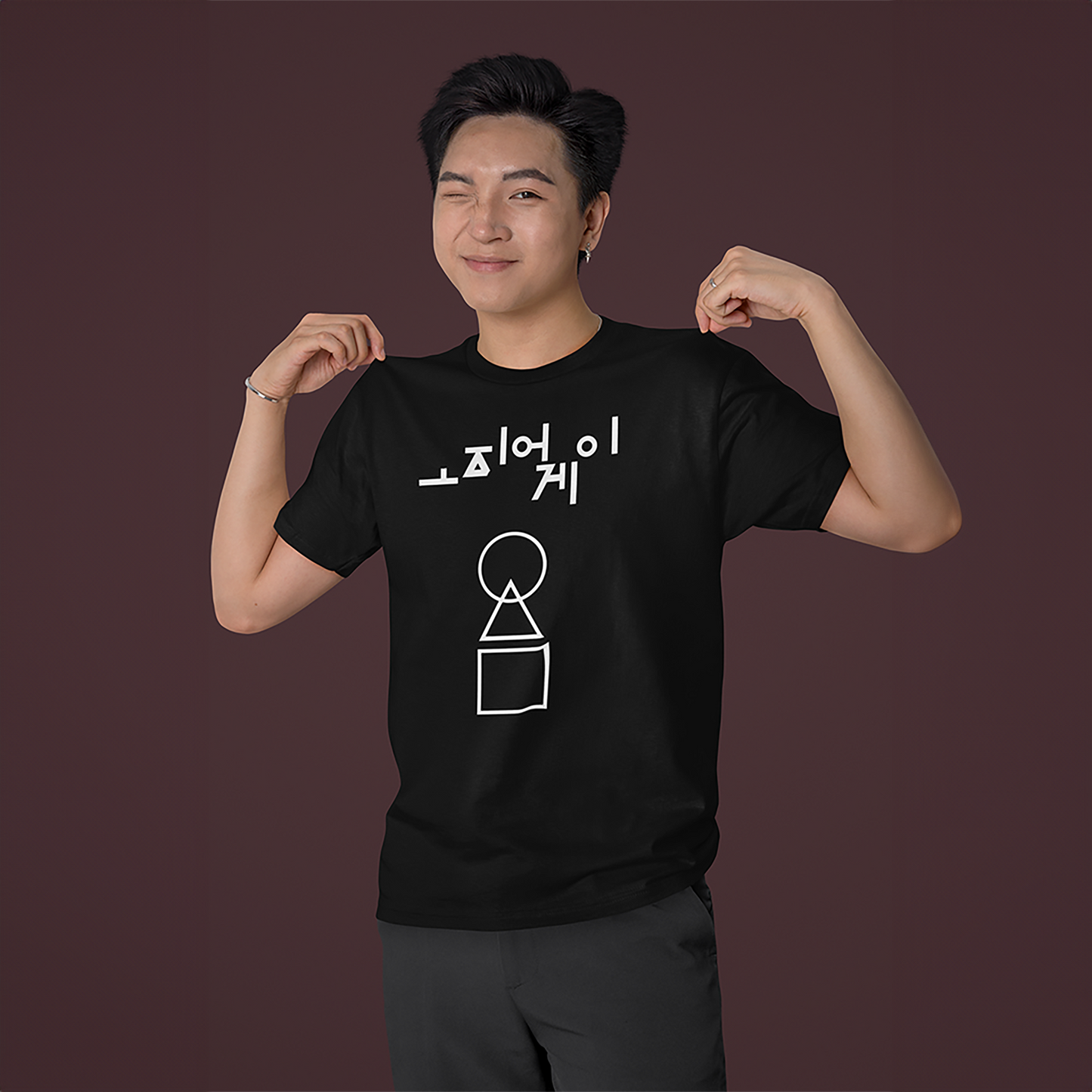 A playful man striking a pose in a black 'Squid Game' T-shirt, showcasing bold white graphics with Korean text and geometric icons. A trendy outfit for casual and fandom-inspired styles. Made by Your iDeas Shop