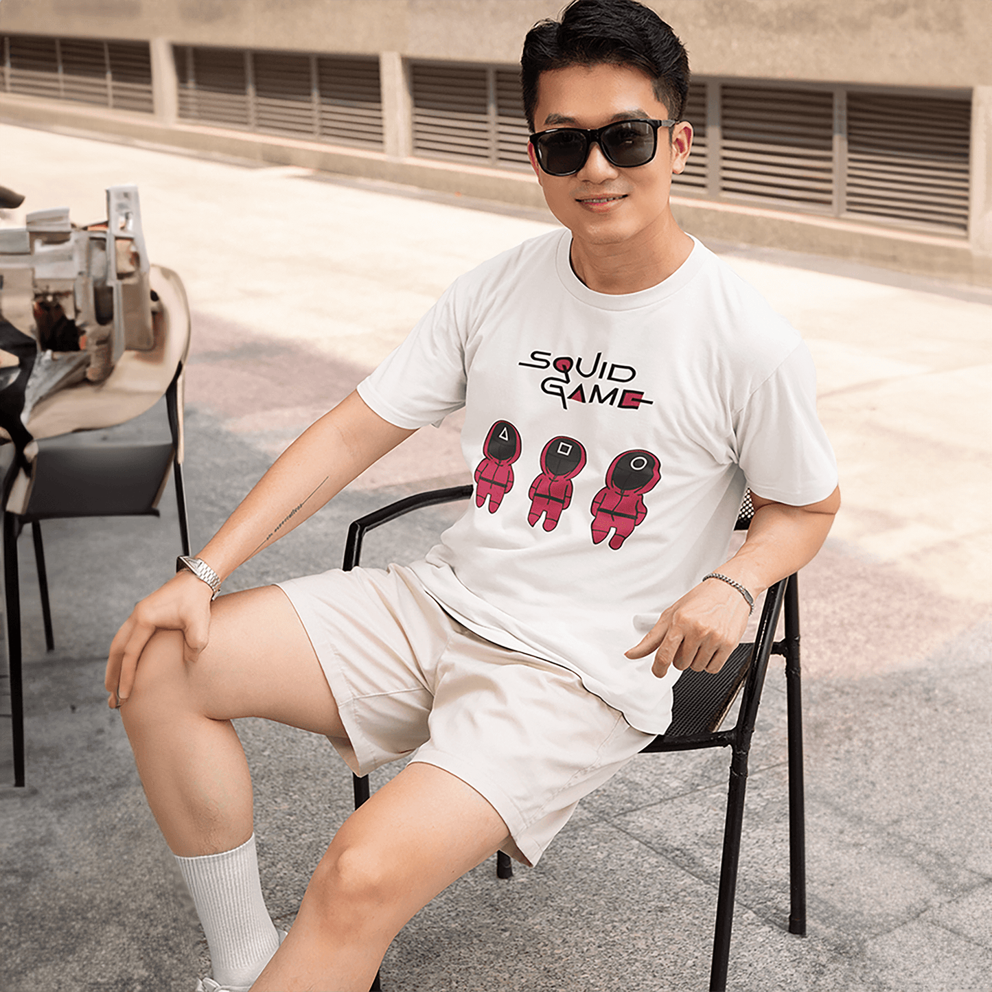 A happy man seated outdoors, wearing a white 'Squid Game' T-shirt with a design of three red cartoon guards and bold text. Ideal for casual outings or relaxed occasions. Made by Your iDeas Shop.