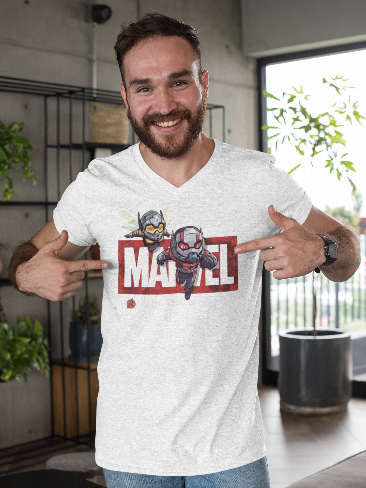 A cheerful man pointing at his white V-neck T-shirt, which displays a chibi-style Ant-Man and Wasp design over a red Marvel logo. Captured in a modern indoor setting. Made by Your iDeas Shop.