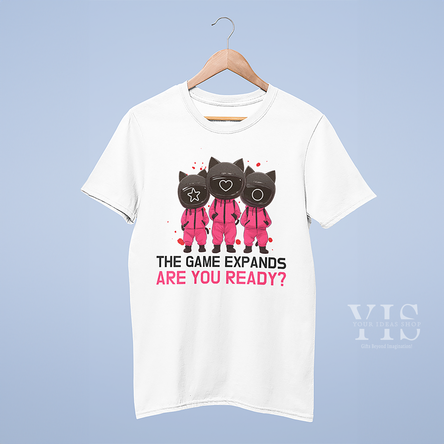 A white T-shirt hanging on a wooden hanger, featuring a fun 'Squid Game' cats graphic with three cartoon cats in pink uniforms, along with the bold phrase 'The Game Expands. Are You Ready?' against a simple blue background. Made by Your iDeas Shop.