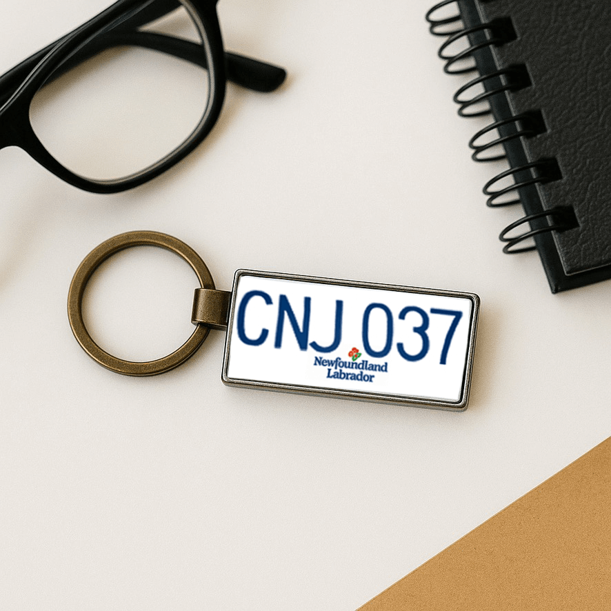 A Newfoundland and Labrador license plate-style keychain with the code 'CNJ 037' displayed on a light desk next to black-rimmed glasses and a spiral notebook. The minimalist composition suggests a smart and professional gift idea with a Canadian touch. Made by Your iDeas Shop.