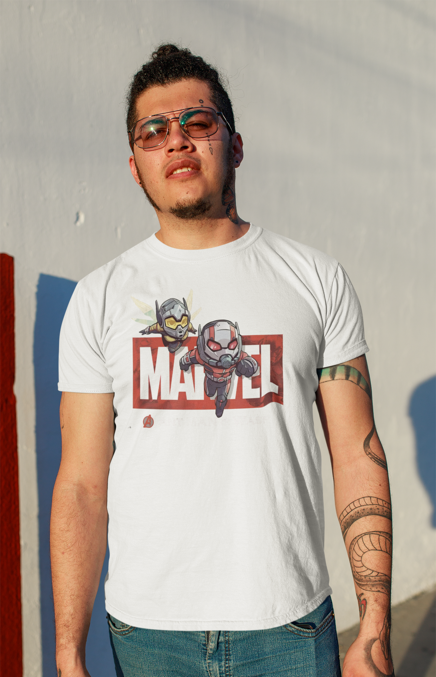 A stylish man with tattoos and sunglasses sporting a white T-shirt with a chibi-style Ant-Man and Wasp print. The urban setting and warm lighting enhance the streetwear vibe. Made by Your iDeas Shop.