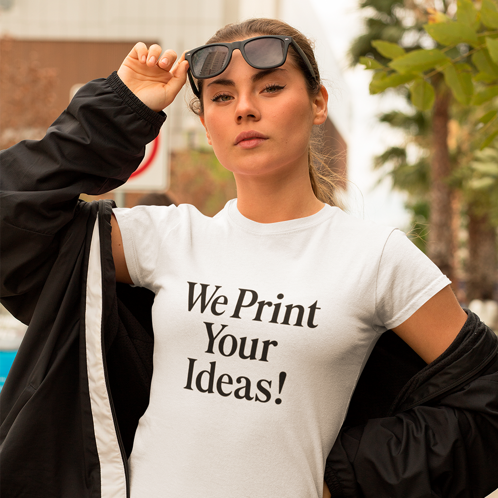 Confident young woman in sunglasses and open jacket showcasing a white T-shirt with bold black slogan ‘We Print Your Ideas!’—perfect for advertising bespoke apparel printing services. Made by Your iDeas Shop