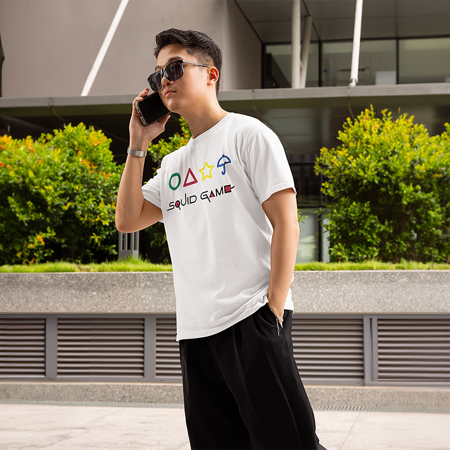 A stylish man wearing a white 'Squid Game' T-shirt, featuring playful geometric icons from the series, speaking on the phone outdoors. Perfect for casual wear. Made by Your iDeas Shop
