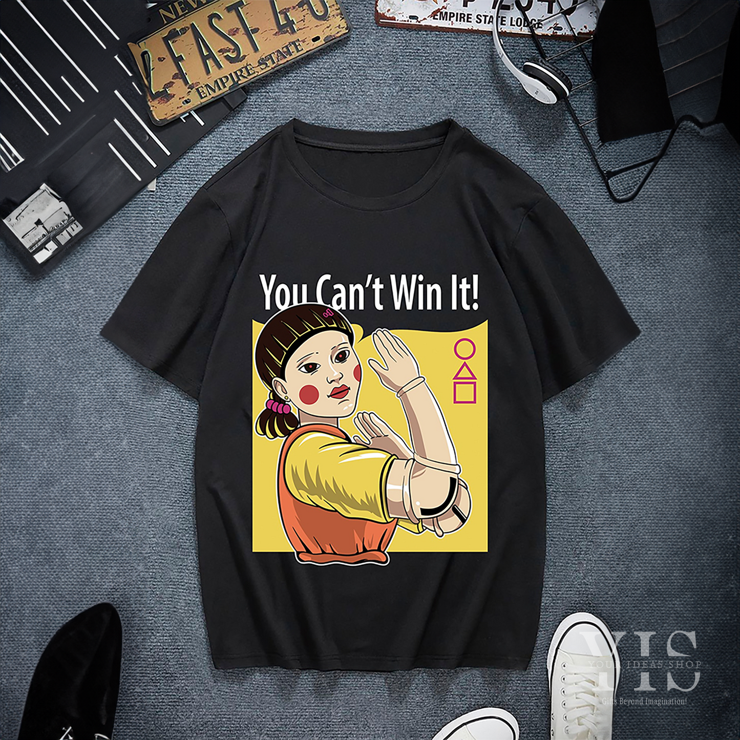 A black T-shirt featuring the iconic 'Red Light, Green Light' doll from Squid Game with the bold text 'You Can't Win It!' in white. The graphic includes a yellow backdrop and geometric symbols, creating a striking and playful design. Made by Your iDeas Shop.
