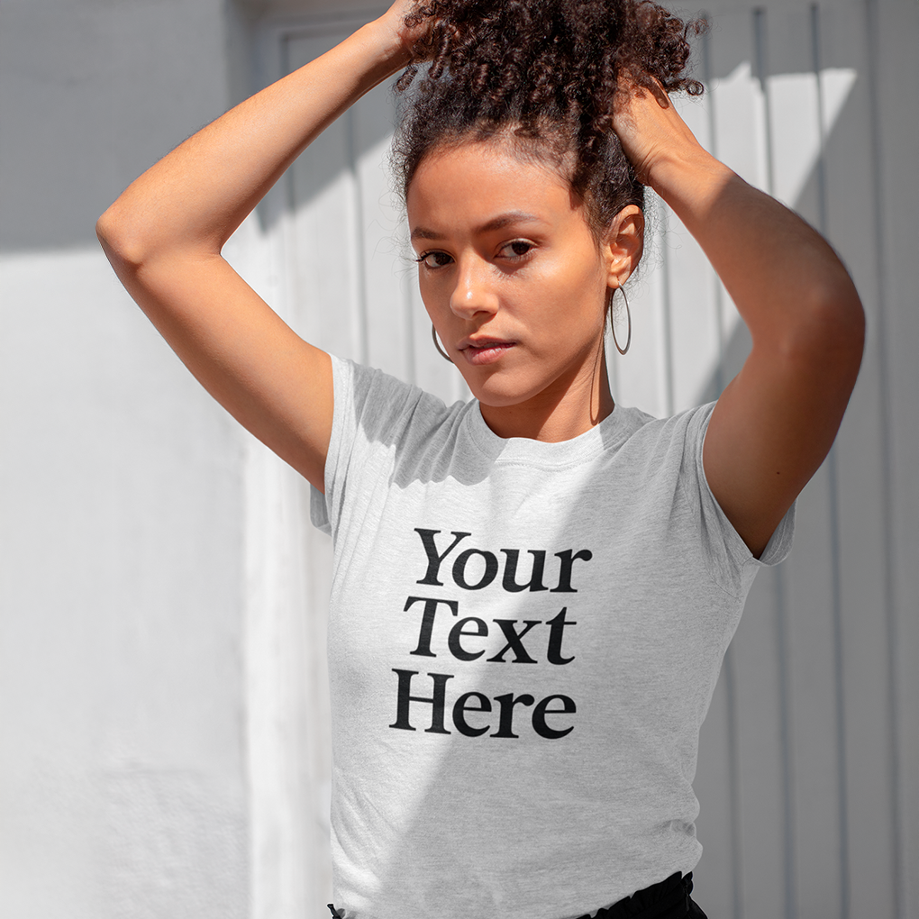 Young woman adjusting her hair in bright sunlight while wearing a light-grey T-shirt that reads ‘Your Text Here’, illustrating a blank template ready for custom wording or graphics. Made by Your iDeas Shop