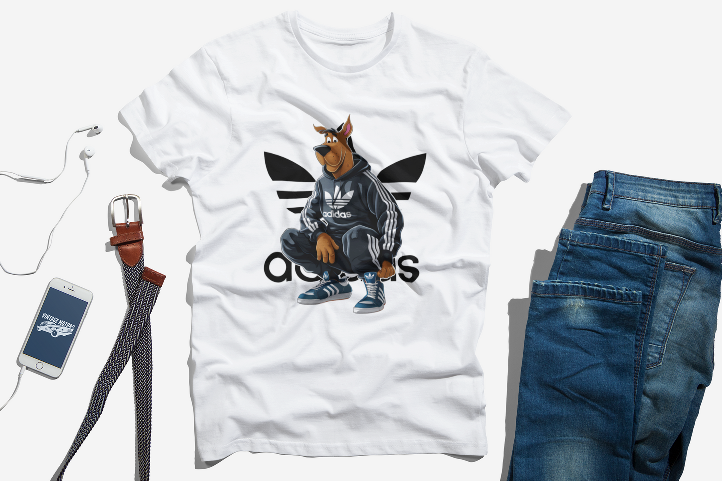 Flat lay of a white t-shirt featuring a cartoon character resembling Scooby-Doo in a black Adidas hoodie, paired with men's accessories including a belt, a smartphone, and blue jeans, arranged on a white background for a casual and stylish outfit look.