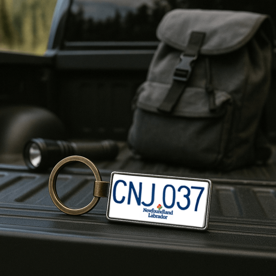A Newfoundland and Labrador license plate-style keychain marked 'CNJ 037' resting on the bed of a pickup truck, with a black backpack and flashlight in the background. This rugged outdoor setting highlights the keychain as a practical and thematic gift for travelers and explorers. Made by Your iDeas Shop.