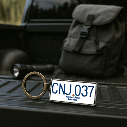 A Newfoundland and Labrador license plate-style keychain marked 'CNJ 037' resting on the bed of a pickup truck, with a black backpack and flashlight in the background. This rugged outdoor setting highlights the keychain as a practical and thematic gift for travelers and explorers. Made by Your iDeas Shop.