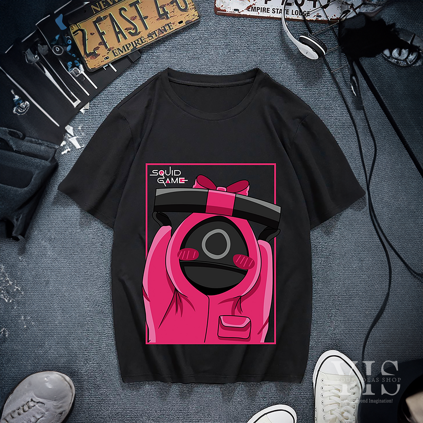 A black T-shirt with a fun cartoon illustration of a pink-uniformed guard holding a gift box. The design includes a pink border and the 'Squid Game' logo for an extra pop. A standout shirt for casual wear or gifting. Made by Your iDeas Shop.