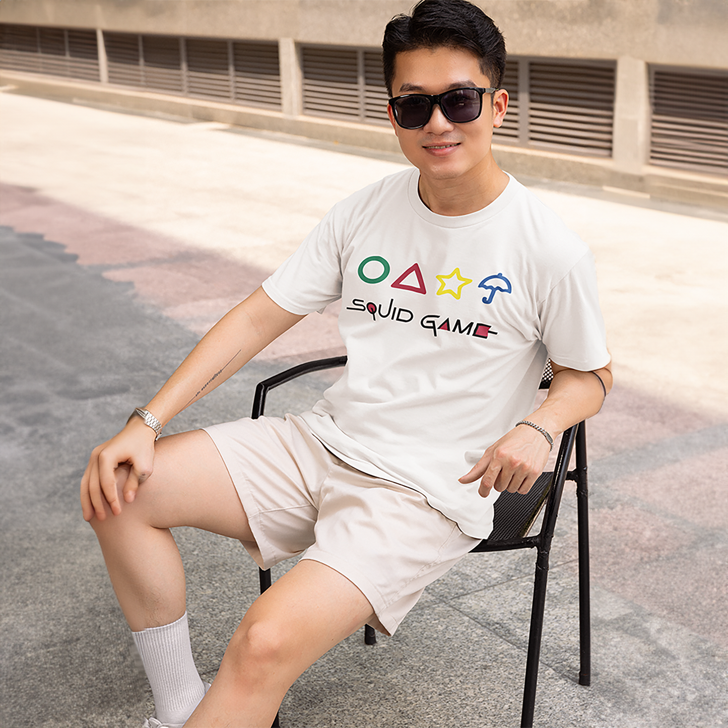 A cheerful man sitting on a chair, wearing a white 'Squid Game' T-shirt with colorful geometric shapes and text. A fun, casual fashion choice. Made by Your iDeas Shop