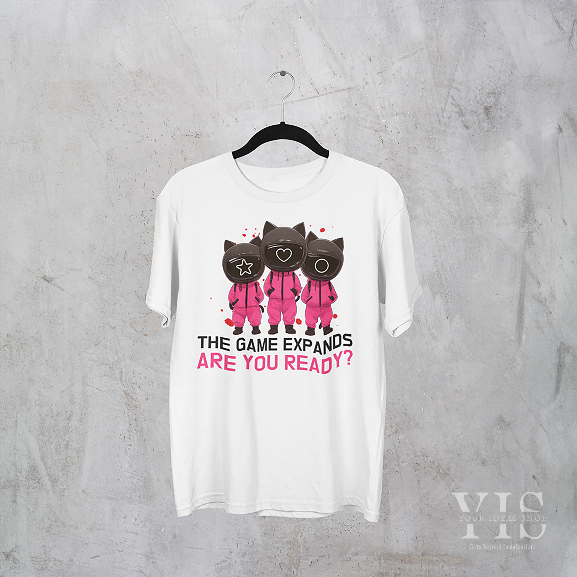 A white T-shirt featuring a playful 'Squid Game' cats design, with three cartoon cats in pink guard uniforms and text reading 'The Game Expands. Are You Ready?' The shirt is displayed against a whimsical space-themed background. Made by Your iDeas Shop