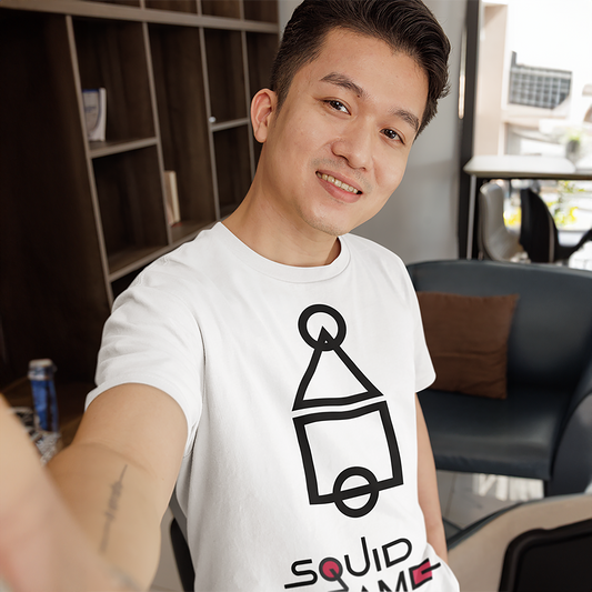 A smiling man taking a selfie indoors, wearing a white 'Squid Game' T-shirt featuring bold geometric icons in black. A modern look for fans of the series. Made by Your iDeas Shop.