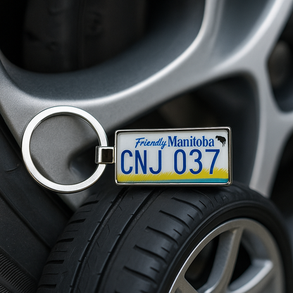 A Friendly Manitoba license plate keychain with CNJ 037 design leaning against a car wheel rim. A perfect travel accessory and gift for road trip enthusiasts. Made by Your iDeas Shop
