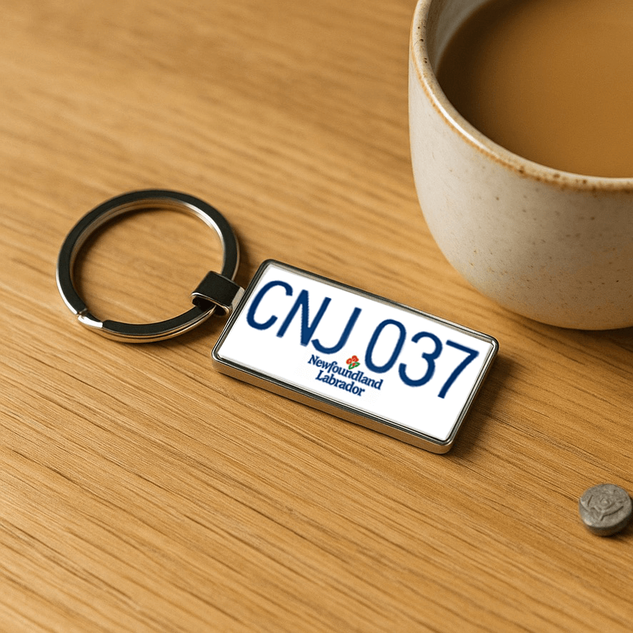 A Newfoundland and Labrador license plate-style keychain featuring the code 'CNJ 037' placed on a light wooden surface next to a ceramic mug filled with coffee. A simple and cozy flatlay, perfect for showcasing everyday accessories or gift ideas. Made by Your iDeas Shop.