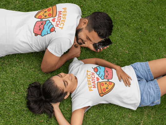 Man and woman relaxing on green grass in coordinated white tees printed with the phrase ‘Perfect Match’ above kawaii pizza and cupcake characters, capturing a playful picnic scene and ideal for romantic gift ideas. Made by Your iDeas Shop