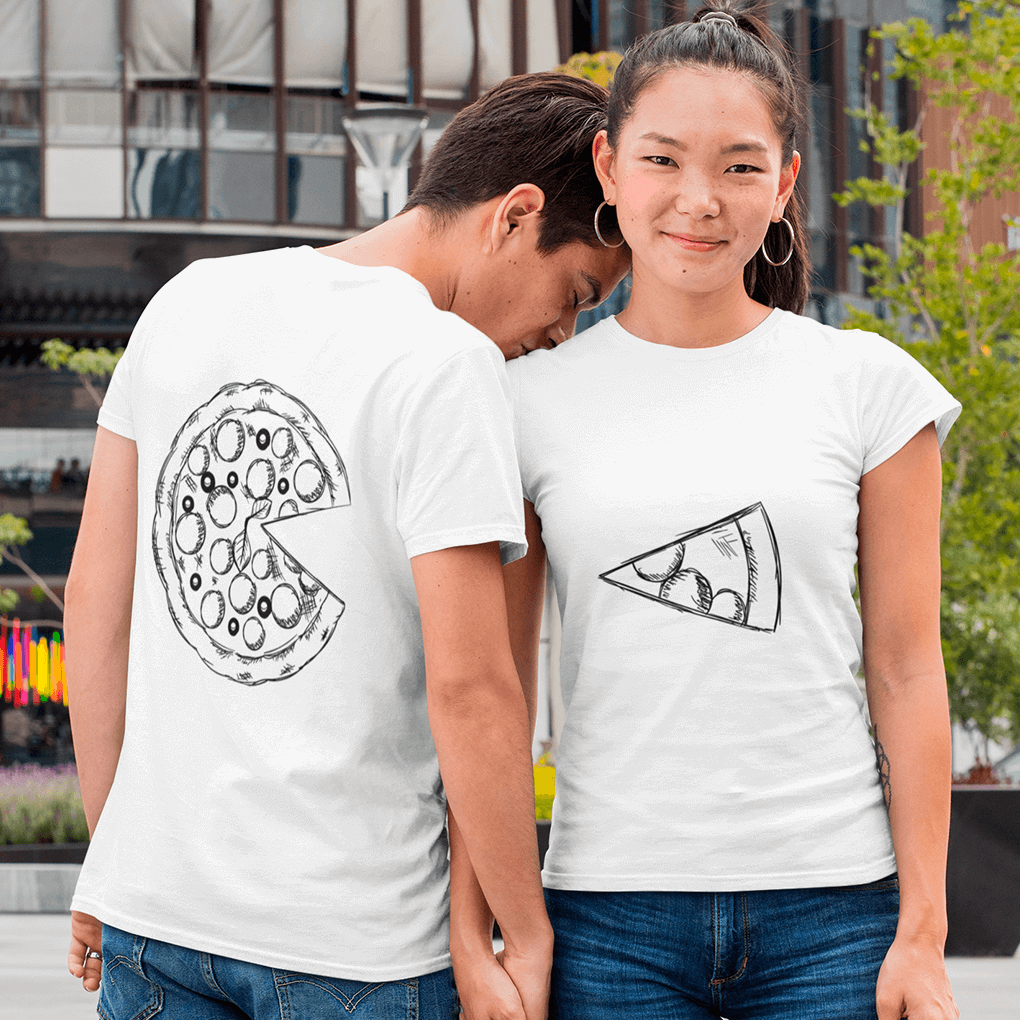 Young couple in casual white matching T-shirts with complementary hand-sketched pizza graphics: his shirt (back view) shows a full pepperoni pizza missing one slice, while hers (front view) displays the single slice. They hold hands and stand in an urban setting, highlighting a playful ‘you complete me’ design for couples. Made by Your iDeas Shop.