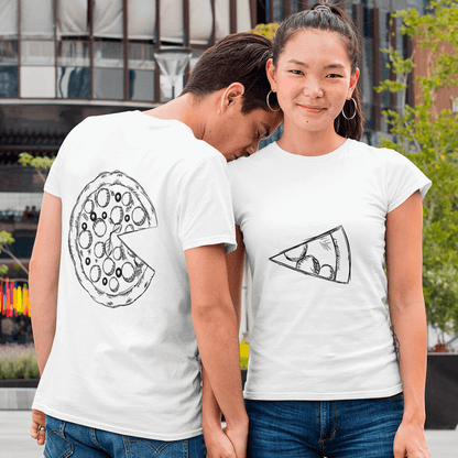Young couple in casual white matching T-shirts with complementary hand-sketched pizza graphics: his shirt (back view) shows a full pepperoni pizza missing one slice, while hers (front view) displays the single slice. They hold hands and stand in an urban setting, highlighting a playful ‘you complete me’ design for couples. Made by Your iDeas Shop.