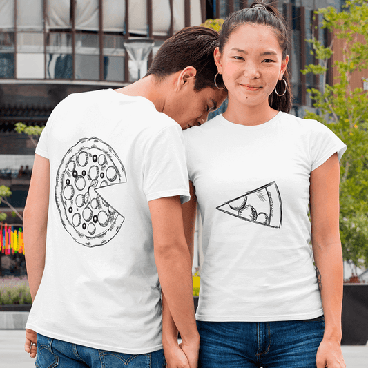 Young couple in casual white matching T-shirts with complementary hand-sketched pizza graphics: his shirt (back view) shows a full pepperoni pizza missing one slice, while hers (front view) displays the single slice. They hold hands and stand in an urban setting, highlighting a playful ‘you complete me’ design for couples. Made by Your iDeas Shop.