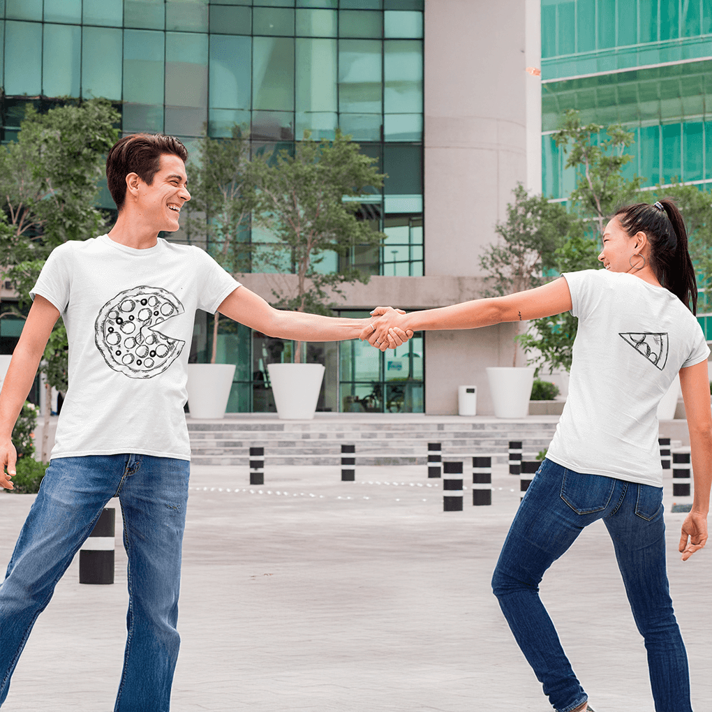 Playful pair stretching arms while holding hands across an open urban plaza, wearing coordinated white shirts—his front printed with the large circular pizza minus one slice, hers (back view) printed with the corresponding slice—symbolizing togetherness for pizza-enthusiast couples. Made by Your iDeas Shop.