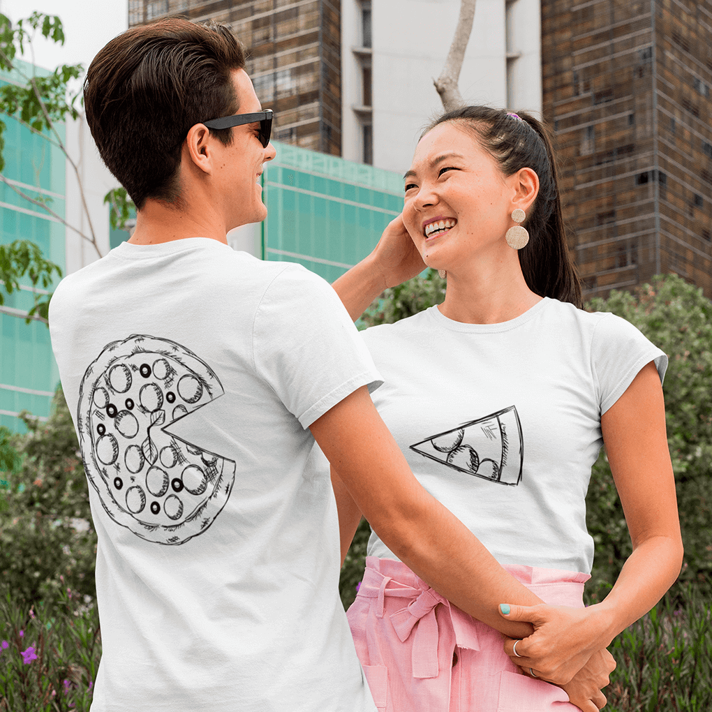 Happy couple laughing together in the city: his white tee (back view) displays a large sketched pizza with a missing slice, while her tee (front view) displays the matching slice graphic, creating a fun complementary look ideal for food-themed gifts. Made by Your iDeas Shop.
