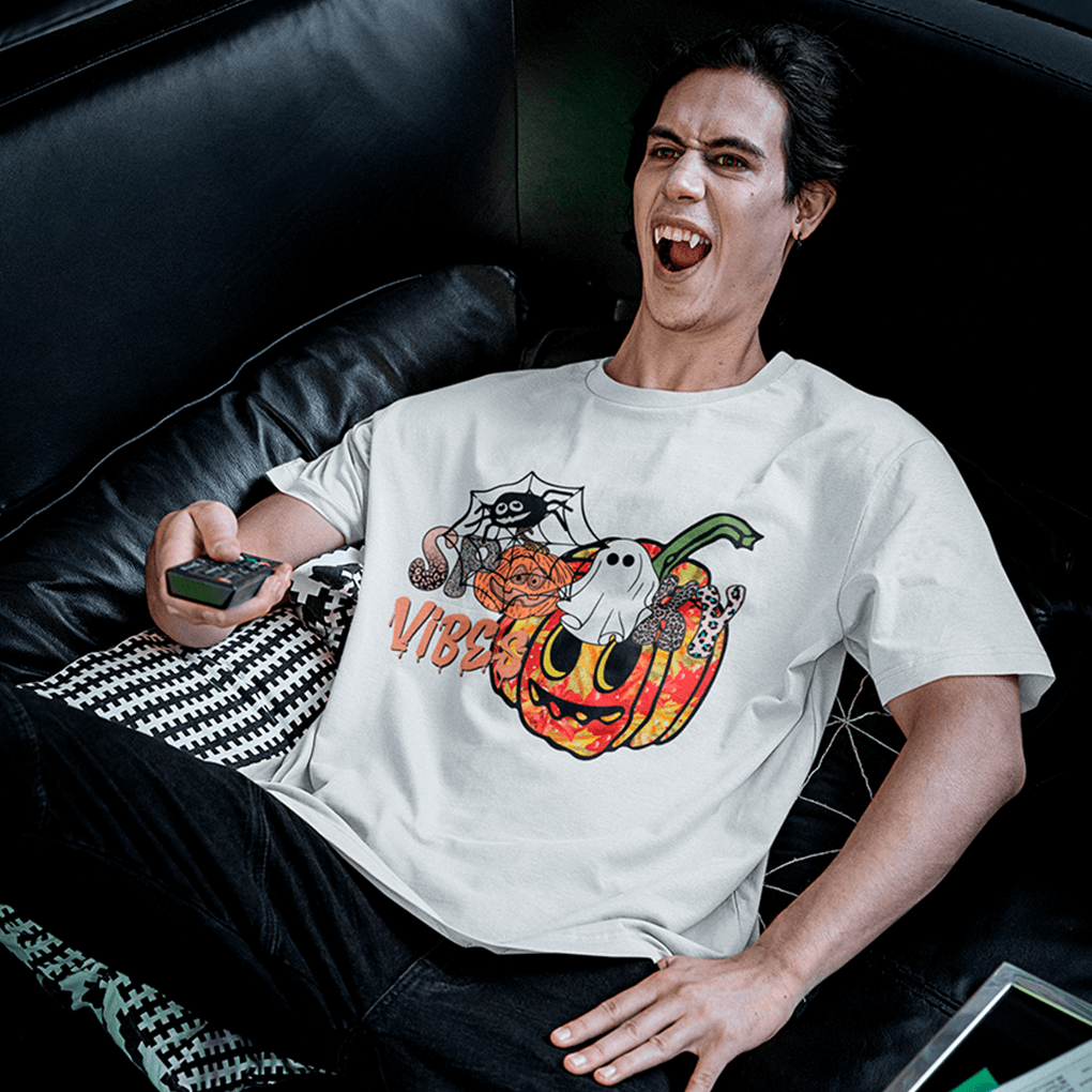 A young man lounging on a black leather sofa, laughing while holding a TV remote, dressed in a white T-shirt that features a bright jack-o’-lantern with cartoon eyes, a cute ghost on the stem, a spider in its web and the dripping phrase ‘Spooky Vibes’—a fun Halloween shirt for horror-movie nights. Made by Your iDeas Shop.