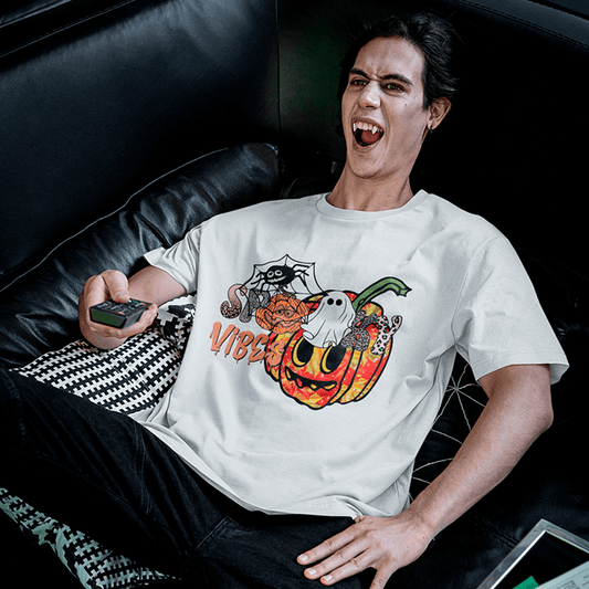 A young man lounging on a black leather sofa, laughing while holding a TV remote, dressed in a white T-shirt that features a bright jack-o’-lantern with cartoon eyes, a cute ghost on the stem, a spider in its web and the dripping phrase ‘Spooky Vibes’—a fun Halloween shirt for horror-movie nights. Made by Your iDeas Shop.