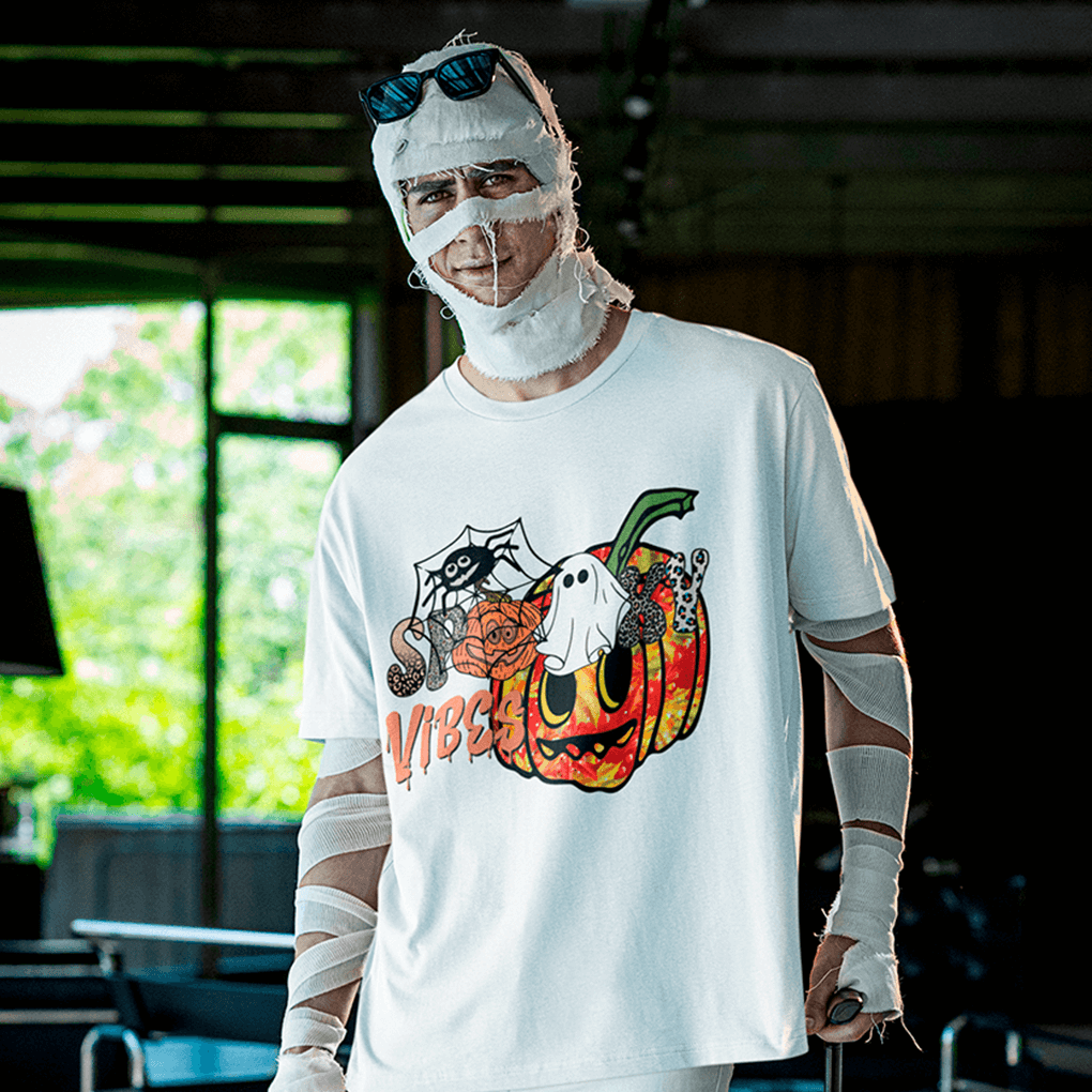 Man wrapped in playful mummy bandages and sunglasses poses indoors wearing a white Halloween T-shirt printed with a colorful grinning pumpkin, cobweb spider, mini ghost and the text ‘Spooky Vibes’, perfect for easy costume parties and October festivities. Made by Your iDeas Shop.