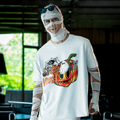 Man wrapped in playful mummy bandages and sunglasses poses indoors wearing a white Halloween T-shirt printed with a colorful grinning pumpkin, cobweb spider, mini ghost and the text ‘Spooky Vibes’, perfect for easy costume parties and October festivities. Made by Your iDeas Shop.