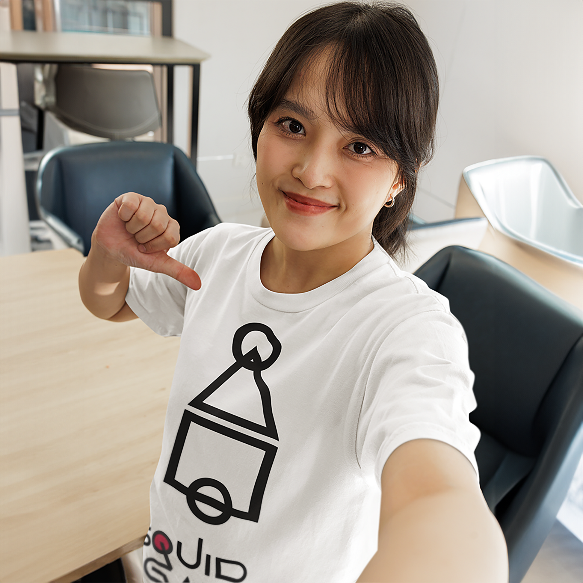 A cheerful woman taking a selfie and pointing at her white 'Squid Game' T-shirt, featuring a bold and minimalist black geometric graphic. A fun and trendy outfit choice. Made by Your iDeas Shop.