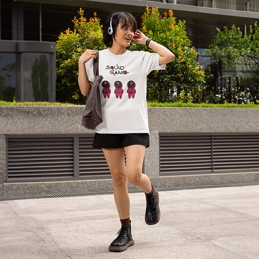 A young woman wearing a white 'Squid Game' T-shirt featuring three red guard characters, listening to music with retro headphones and carrying a tote bag outdoors. A fun and casual outfit idea. Made by Your iDeas ShopA young woman wearing a white 'Squid Game' T-shirt featuring three red guard characters, listening to music with retro headphones and carrying a tote bag outdoors. A fun and casual outfit idea. Made by Your iDeas Shop