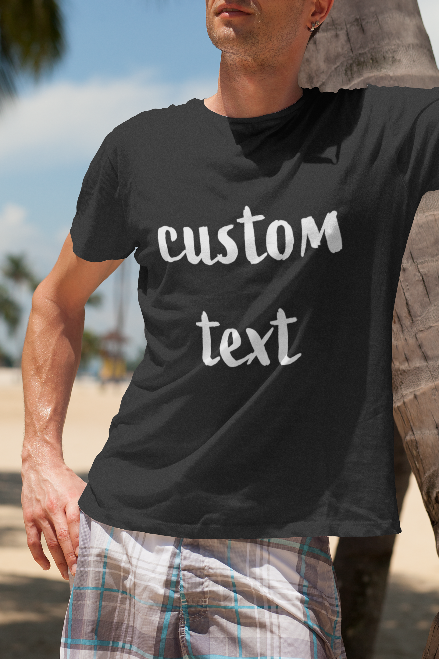 A man leaning against a palm tree while wearing a black t-shirt with the text 'custom text' printed on it. He is standing on a beach, with blurred palm trees and a sunny sky in the background, giving a relaxed and casual vibe. Created by Your iDeas Shop.